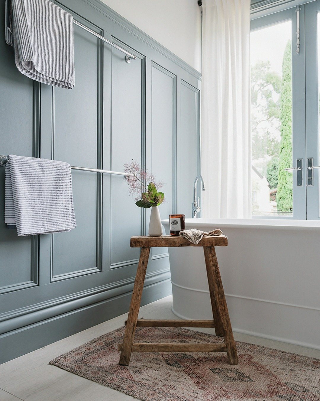 Blue-grey wainscoting panels on a bathroom wall next to a white bathtub and wooden stool.