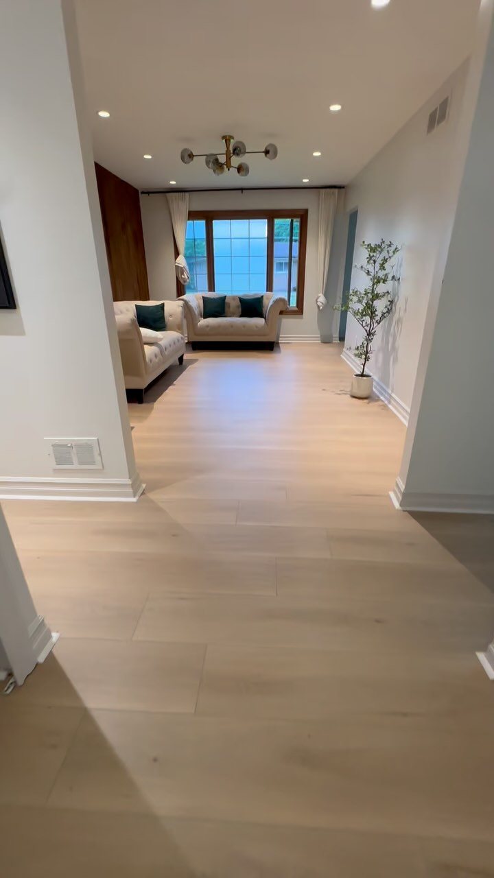 Light wood plank flooring leading into a bright living room with a beige sofa and large windows.