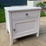 White wooden bedside table with a drawer and cupboard on a stone patio.