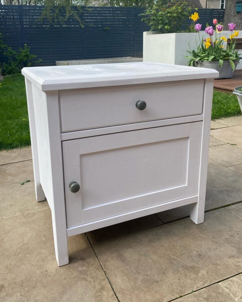 White wooden bedside table with a drawer and cupboard on a stone patio.