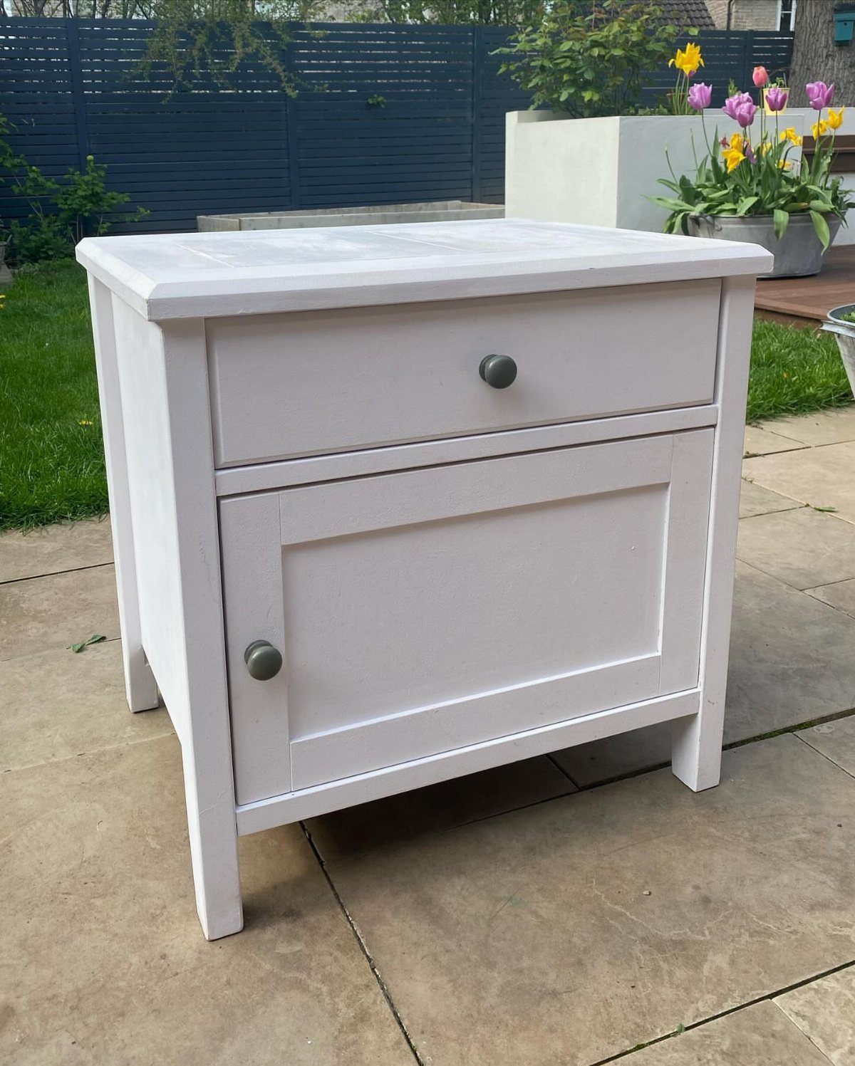 White wooden bedside table with a drawer and cupboard on a stone patio.