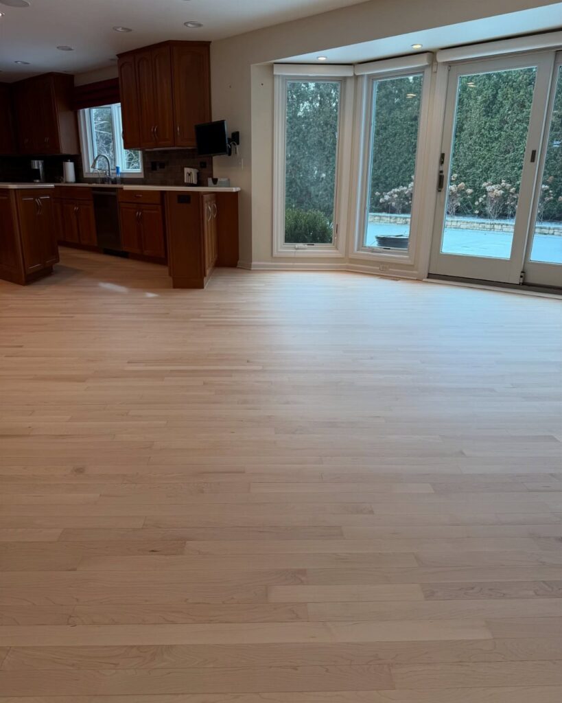 Light maple wood flooring spanning across a large, bright room with kitchen cabinets and glass doors.