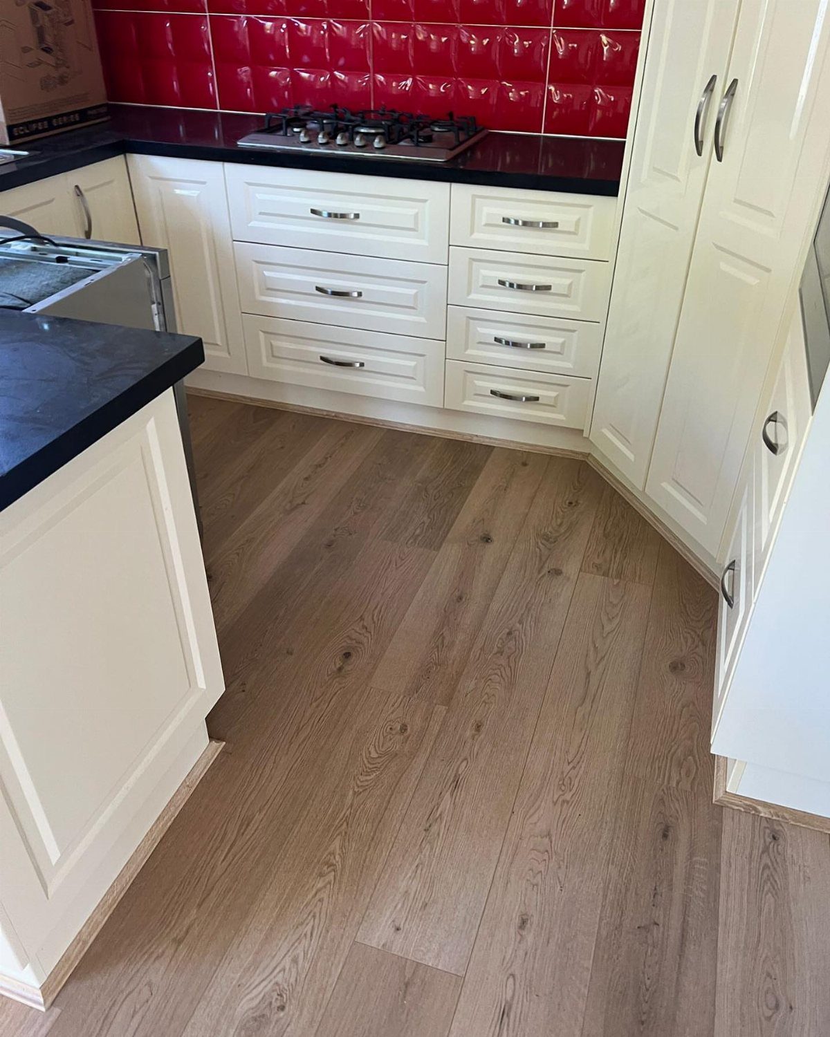 Light oak wood flooring in a kitchen with white cabinets and red backsplash.