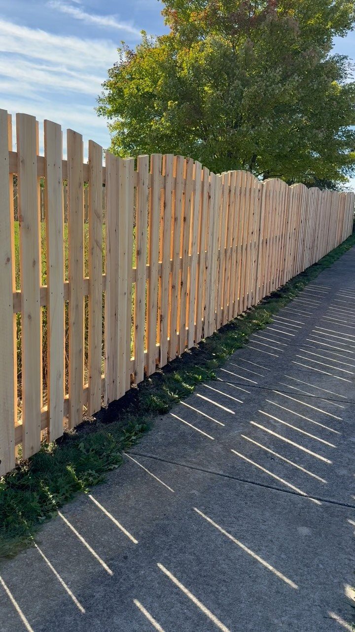 A long wooden shadowbox fence running alongside a concrete sidewalk with sunlight casting striped shadows.