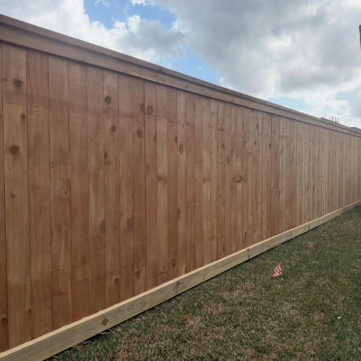 A tall wooden fence with vertical boards stands next to a green grass lawn under a cloudy sky.