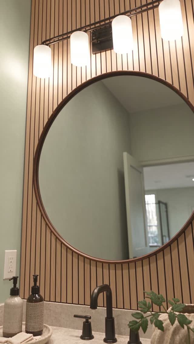 Vertical wooden slats on a bathroom wall behind a large round mirror and vanity.
