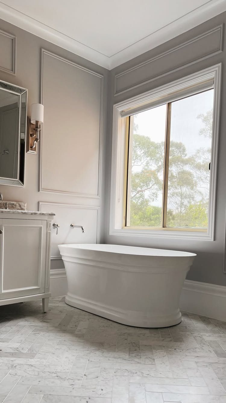 White freestanding bathtub positioned against grey decorative wainscoted walls near a large window.