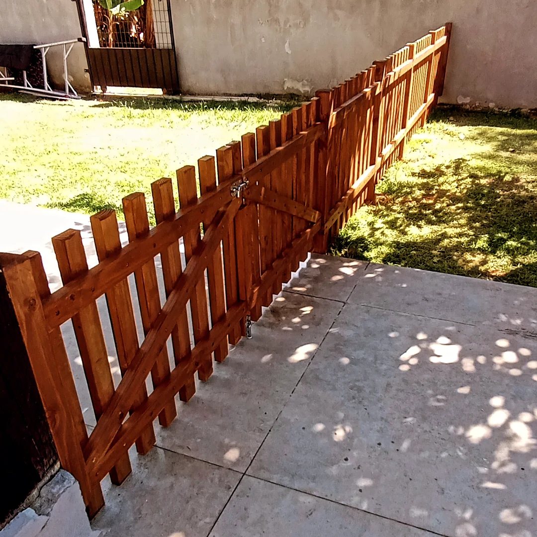 A brown wooden picket fence with a matching gate installed along a concrete patio and grass.
